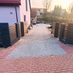 Ułożona z kostki brukowej, dwukolorowa droga dojazdowa do domu z nowoczesnym ogrodzeniem panelowym i widoczną instalacją elektryczną pod ziemią.