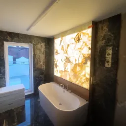 Nowoczesna łazienka z podświetlaną ścianą z onyksu, wolnostojąca wanna, ciemna marmurowa okładzina ścian i podłogi, oraz stos białych płyt przy oknie.