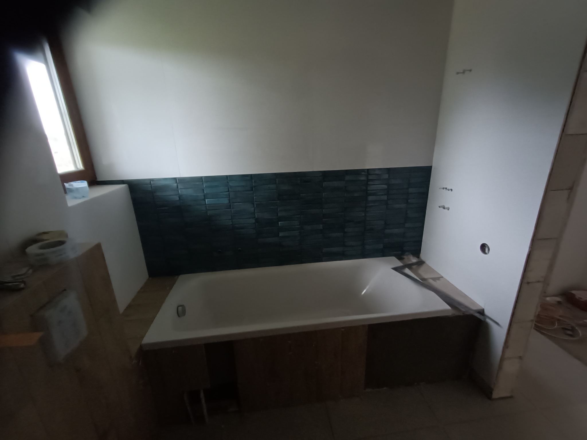 Biała wanna obudowana drewnopodobnymi panelami, z fragmentem ściany wyłożonej ciemnozieloną, strukturalną glazurą w trakcie montażu, widoczne narzędzia i niedokończone prace wykończeniowe.