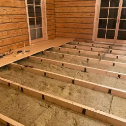 Wnętrze drewnianego domu w trakcie prac: izolacja podłogi wełną mineralną, częściowo ułożone deski podłogowe, widoczne narzędzia.