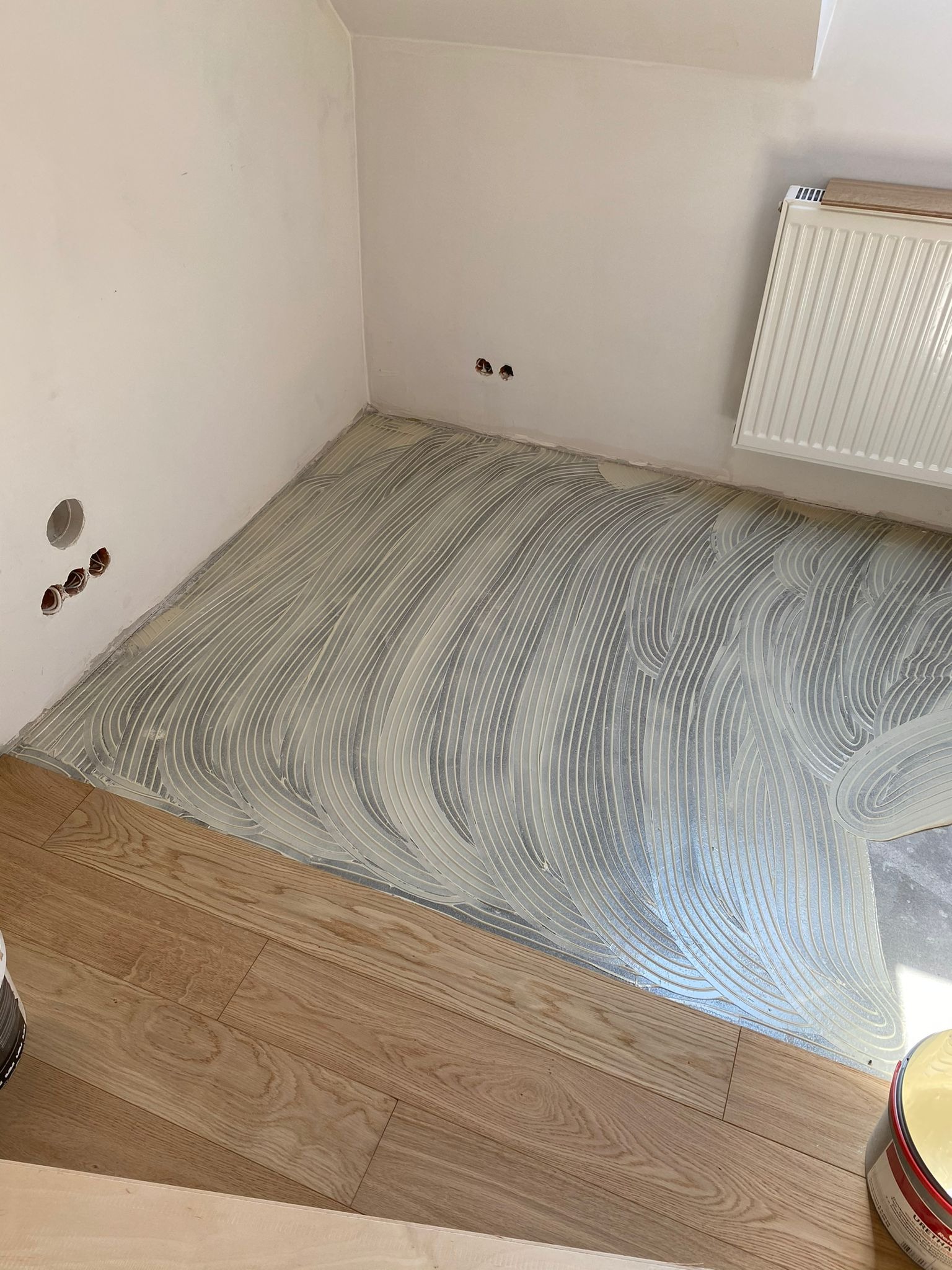 Podłoże przygotowane klejem w charakterystyczne fale pod montaż jasnego parkietu w trakcie prac wykończeniowych w pomieszczeniu z grzejnikiem.