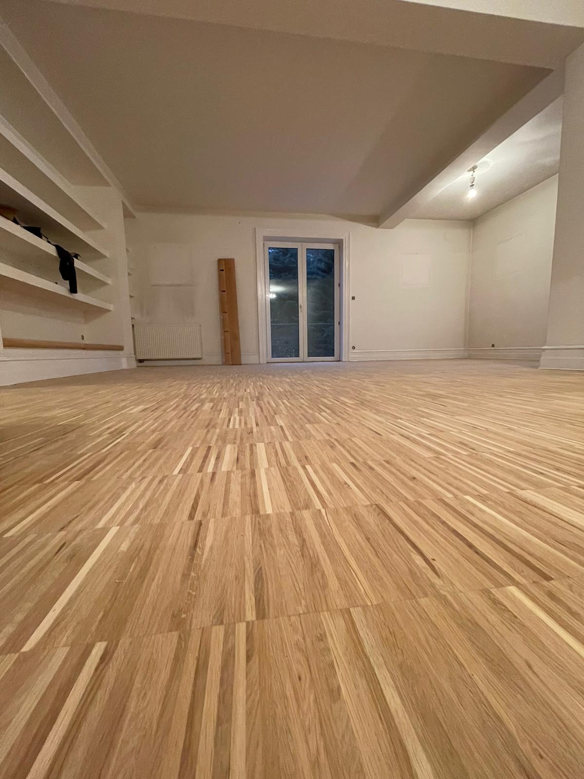 Nowo ułożony, jasny parkiet w pustym pokoju z białymi ścianami, widoczne drzwi balkonowe i wbudowane półki.