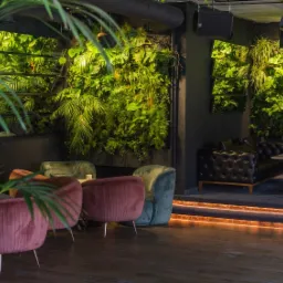 Wnętrze klubu z żywą ścianą z roślin, welurowe fotele w odcieniach różu i zieleni, skórzane sofy Chesterfield oraz podświetlane schody.