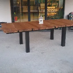 Stół z grubych desek o nieregularnych krawędziach, na czarnych, metalowych nogach, ustawiony na zewnątrz, z ozdobnym lampionem na blacie.