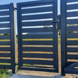 Nowoczesna brama wjazdowa z poziomych, metalowych profili w kolorze grafitowym, z widocznymi elementami mocującymi, na tle zieleni i błękitnego nieba.