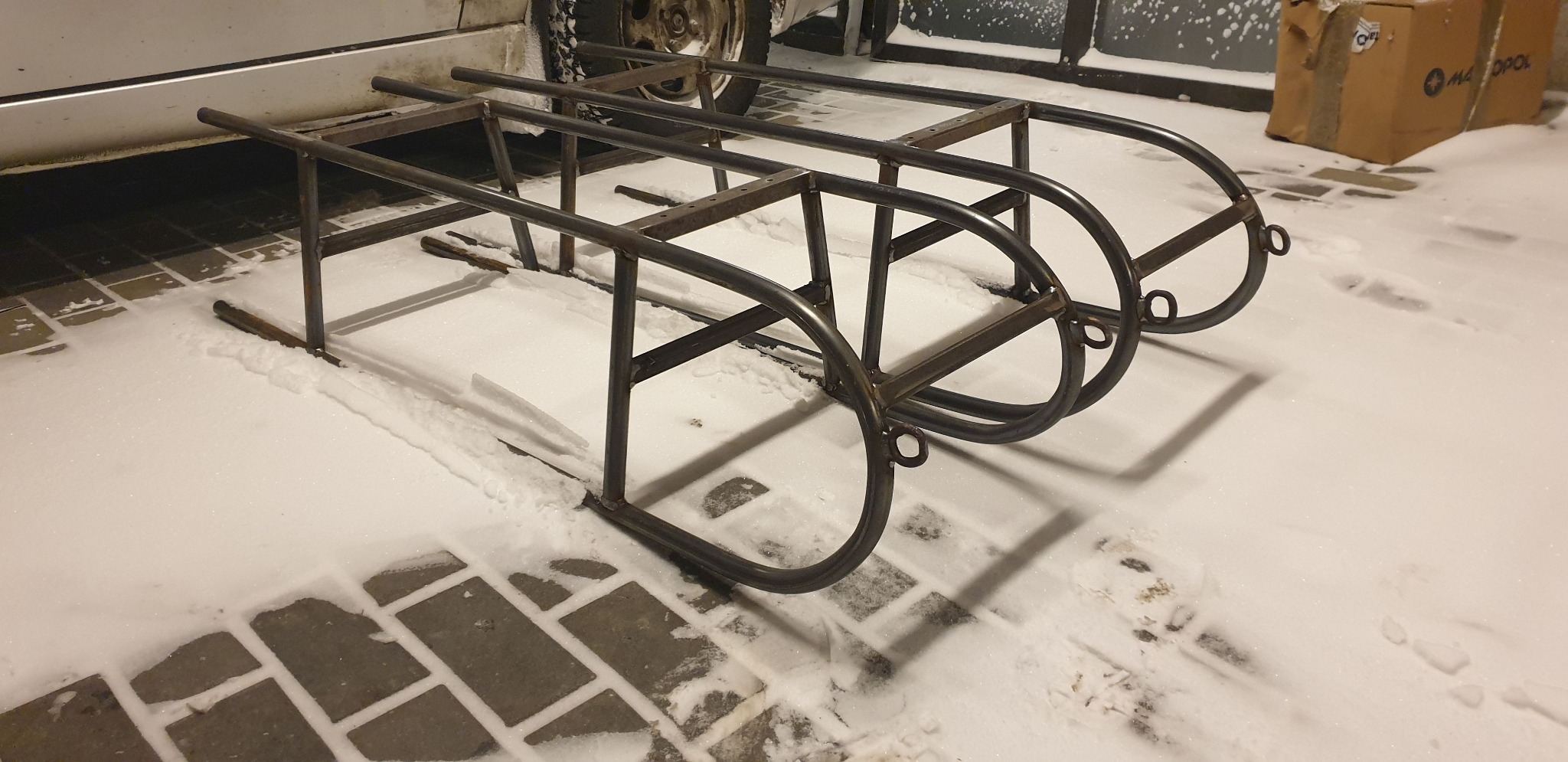 Surowa, metalowa konstrukcja trójsegmentowego wózka transportowego, spawana z rur, stojąca na kostce brukowej pokrytej śniegiem, częściowo pod zaparkowanym samochodem.