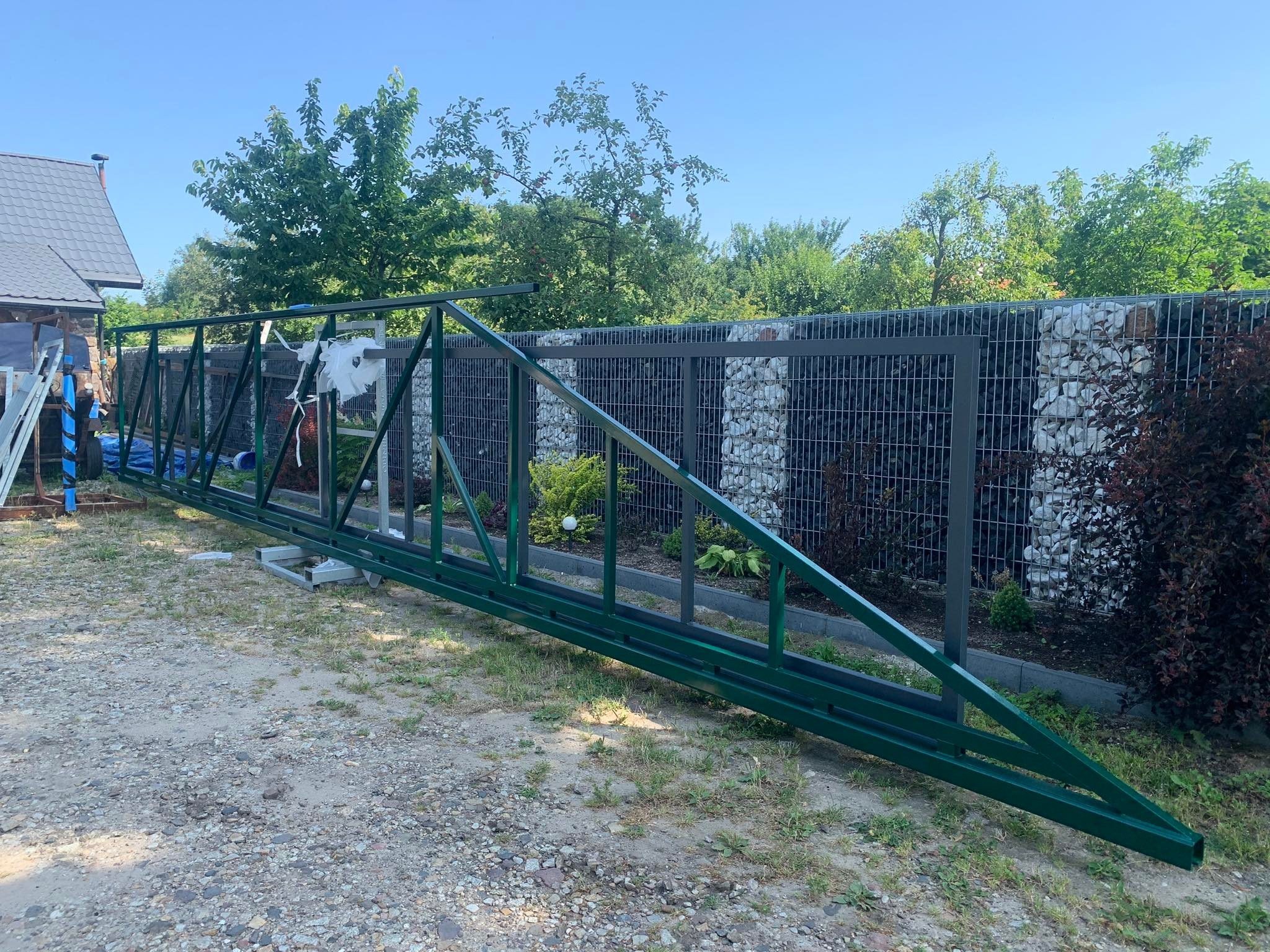Zielona, metalowa konstrukcja bramy przesuwnej leżąca na ziemi obok gabionowego ogrodzenia z białych kamieni, w tle dom z szarą dachówką i drzewa.
