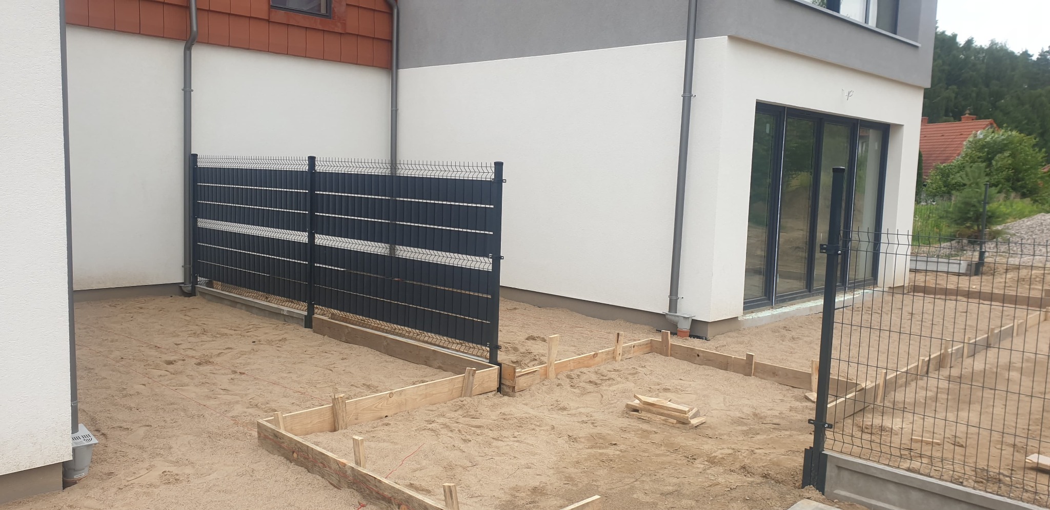 Częściowo zamontowane czarne ogrodzenie panelowe z drewnianym szalunkiem pod wylewkę fundamentu w piasku przy nowoczesnym domu z szarymi elementami.