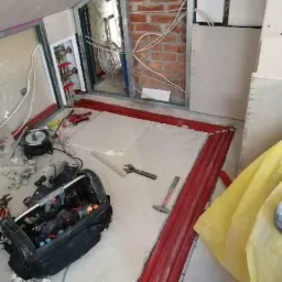 Wnętrze w trakcie instalacji ogrzewania podłogowego: czerwone rury grzewcze ułożone wzdłuż ściany, skrzynka z narzędziami, manometr, ściany w trakcie wykończenia. Widoczny fragment instalacji...