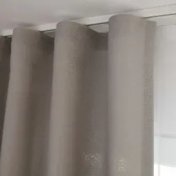Ułożone fałdy szaro-beżowej, lnianej zasłony na metalowym karniszu, z widoczną białą roletą w tle.