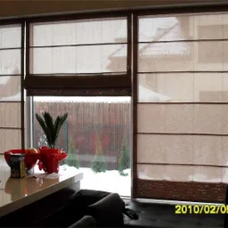 Trzy okna z brązowymi roletami w stylu rzymskim, częściowo podniesionymi; za oknem widoczny ośnieżony ogród z bambusowym płotem; na pierwszym planie blat z owocami w czerwonej misie i doniczką...