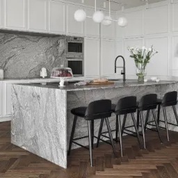 Nowoczesna kuchnia z wyspą wykończoną szarym kamieniem o wyraźnym wzorze, cztery czarne hokery, parkiet w jodełkę, białe szafki i lampa sufitowa z kulistych kloszy. Na blacie chleb na desce, owoce...