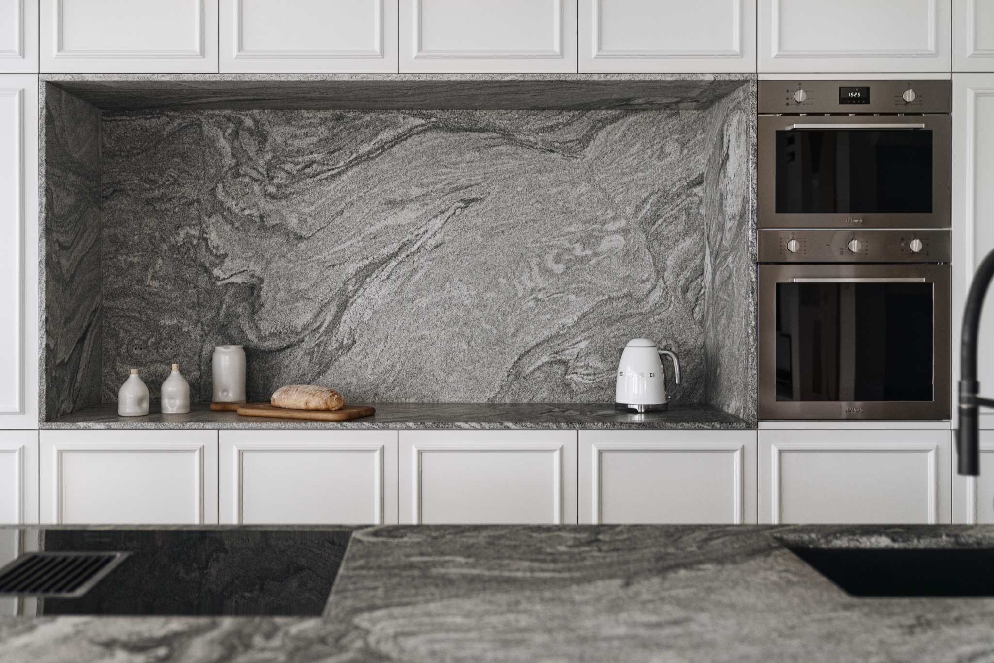 Nowoczesna kuchnia z szarym kamieniem na ścianie i blacie, białe szafki, piekarnik w zabudowie i biały czajnik SMEG.