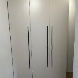 Trzydrzwiowa szafa w kolorze złamanej bieli z długimi, czarnymi uchwytami, widoczna na tle parkietu ułożonego w jodełkę.