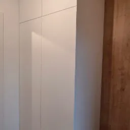 Minimalistyczna, biała szafa wnękowa z gładkimi frontami, zamontowana od podłogi do sufitu w Elblągu, z widoczną drewnianą okładziną ściany po prawej stronie.