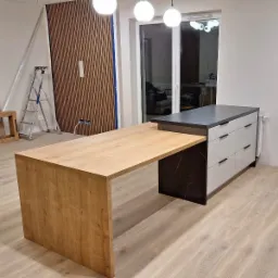 Wnętrze w trakcie wykończenia: drewniany blat roboczy połączony z szafką z szufladami i blatem z ciemnego marmuru, w tle lamelowa ściana i drabina.