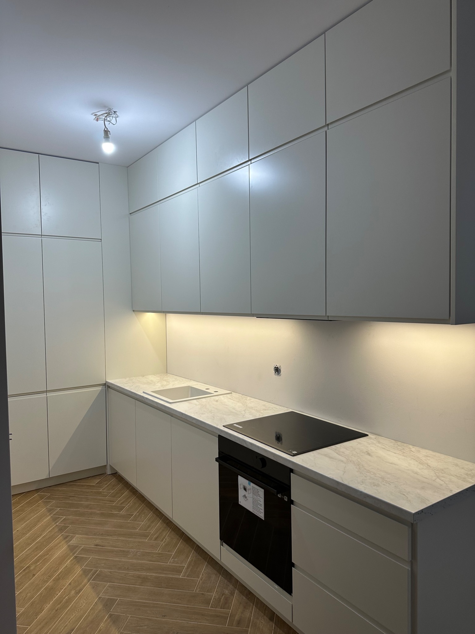 Nowoczesna kuchnia z białymi, matowymi frontami szafek, blatem imitującym marmur i drewnianą podłogą ułożoną w jodełkę. Pod szafkami górnymi zamontowane oświetlenie LED.