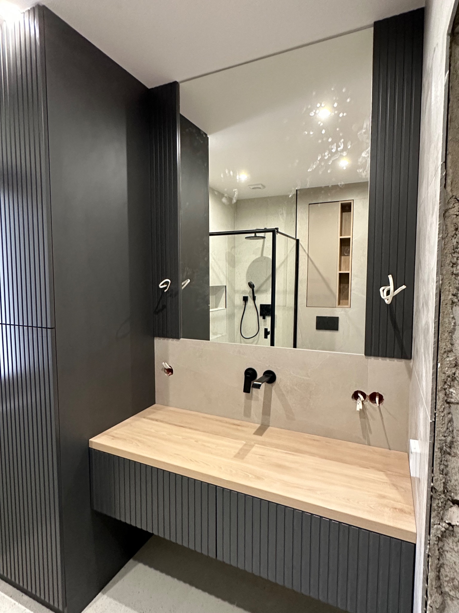 Nowoczesna łazienka w trakcie wykończenia: drewniany blat umywalki na szafce z ryflowanymi frontami, czarna armatura, lustro z widokiem na kabinę prysznicową i wnękę z półkami, widoczne przewody...
