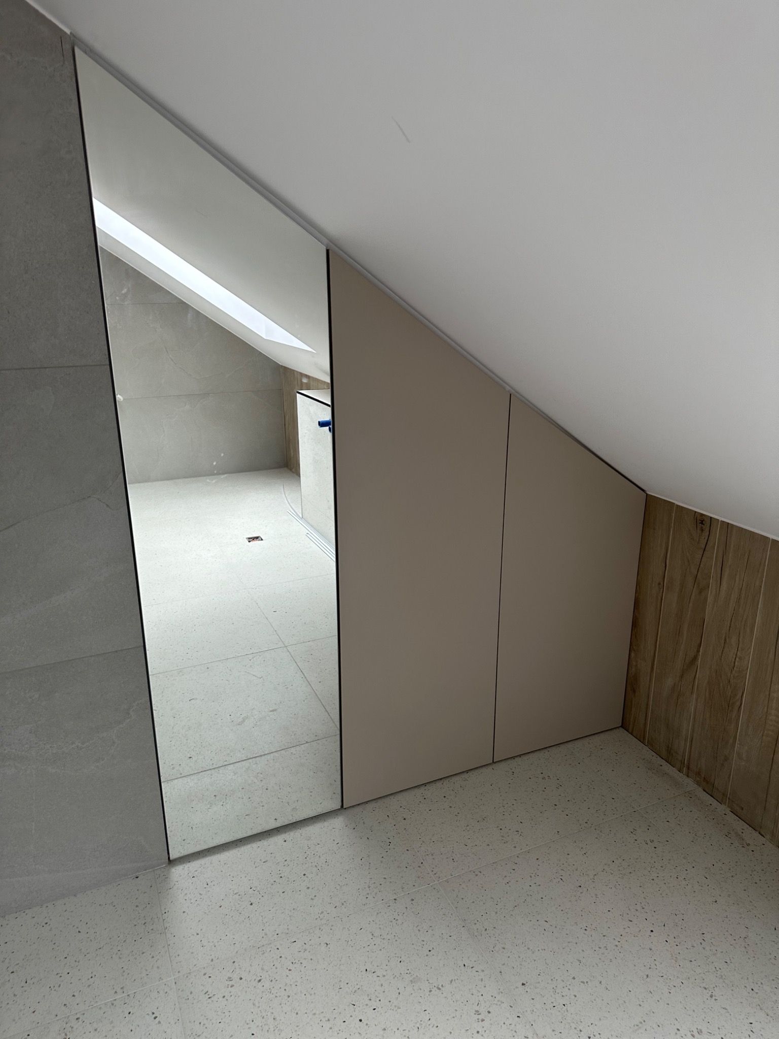 Beżowa szafa wnękowa pod skosem dachu, z lustrzanym frontem, odbijająca wnętrze łazienki z jasnymi płytkami podłogowymi i ściennymi.