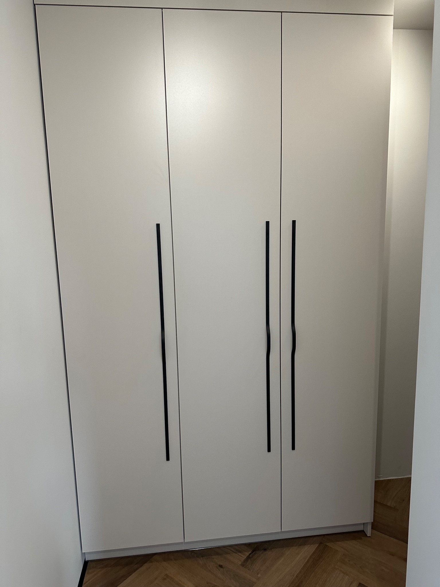 Trzydrzwiowa szafa w kolorze złamanej bieli z długimi, czarnymi uchwytami, widoczna na tle parkietu ułożonego w jodełkę.