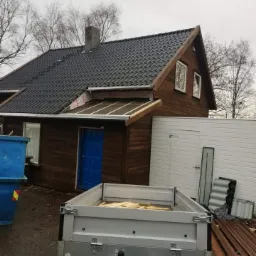 Dom z ciemnobrązową elewacją i nowym, ciemnym dachem z dachówki, przyczepka z drewnem na podwórku, widoczne prace dekarskie w toku.