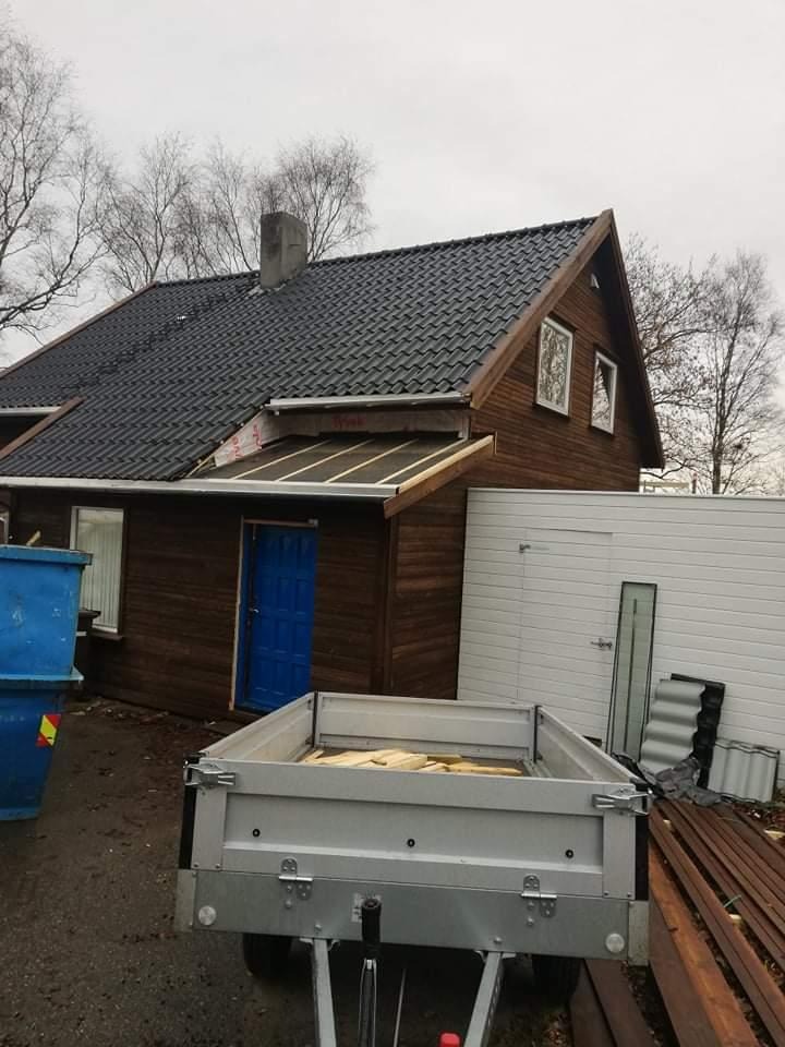 Dom z ciemnobrązową elewacją i nowym, ciemnym dachem z dachówki, przyczepka z drewnem na podwórku, widoczne prace dekarskie w toku.