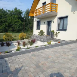 Nowoczesny dom z jasną elewacją, balkonem z drewnianą balustradą i podjazdem wyłożonym kostką w odcieniach szarości i beżu, ozdobnym ogrodem z białym kamieniem i roślinami.