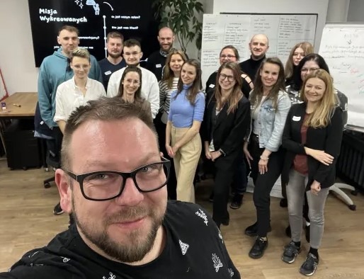 Selfie mężczyzny w okularach na tle zespołu podczas burzy mózgów, z tablicami i notatkami w tle.