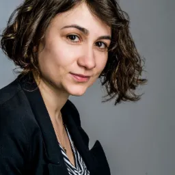 Portret kobiety z brązowymi kręconymi włosami, ubranej w czarną marynarkę i bluzkę w czarno-białe paski, na szarym tle.