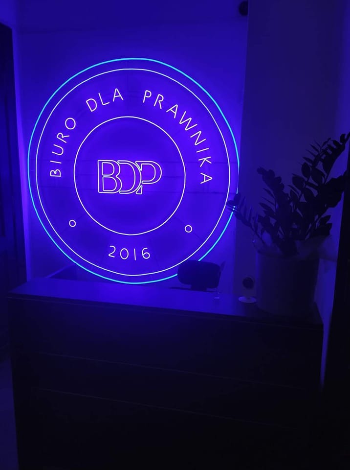 Podświetlany neon w kolorze niebieskim z napisem 'Biuro Dla Prawnika BDP 2016', umieszczony nad ciemnym biurkiem, obok rośliny doniczkowej.