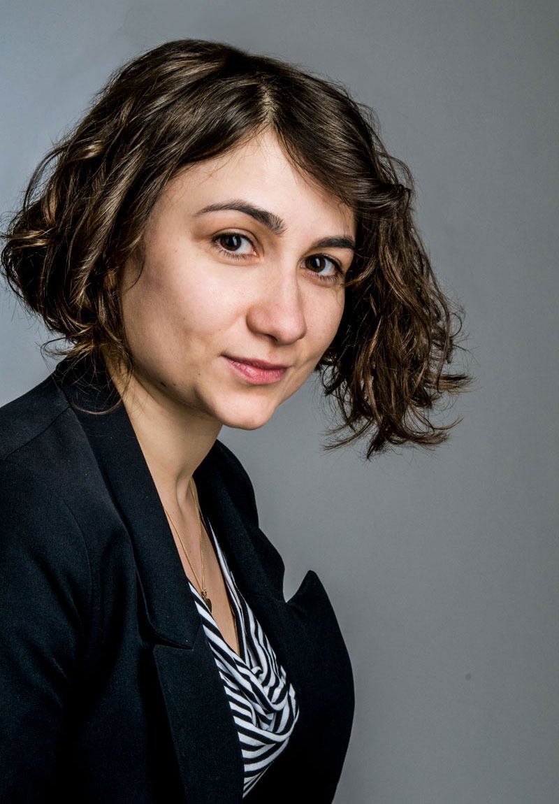 Portret kobiety z brązowymi kręconymi włosami, ubranej w czarną marynarkę i bluzkę w czarno-białe paski, na szarym tle.