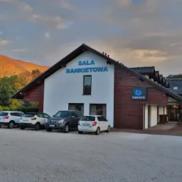 Budynek z napisem 'SALA BANKIETOWA' i logo 'ONESTO' na tle jesiennego krajobrazu górskiego oraz zaparkowane samochody.
