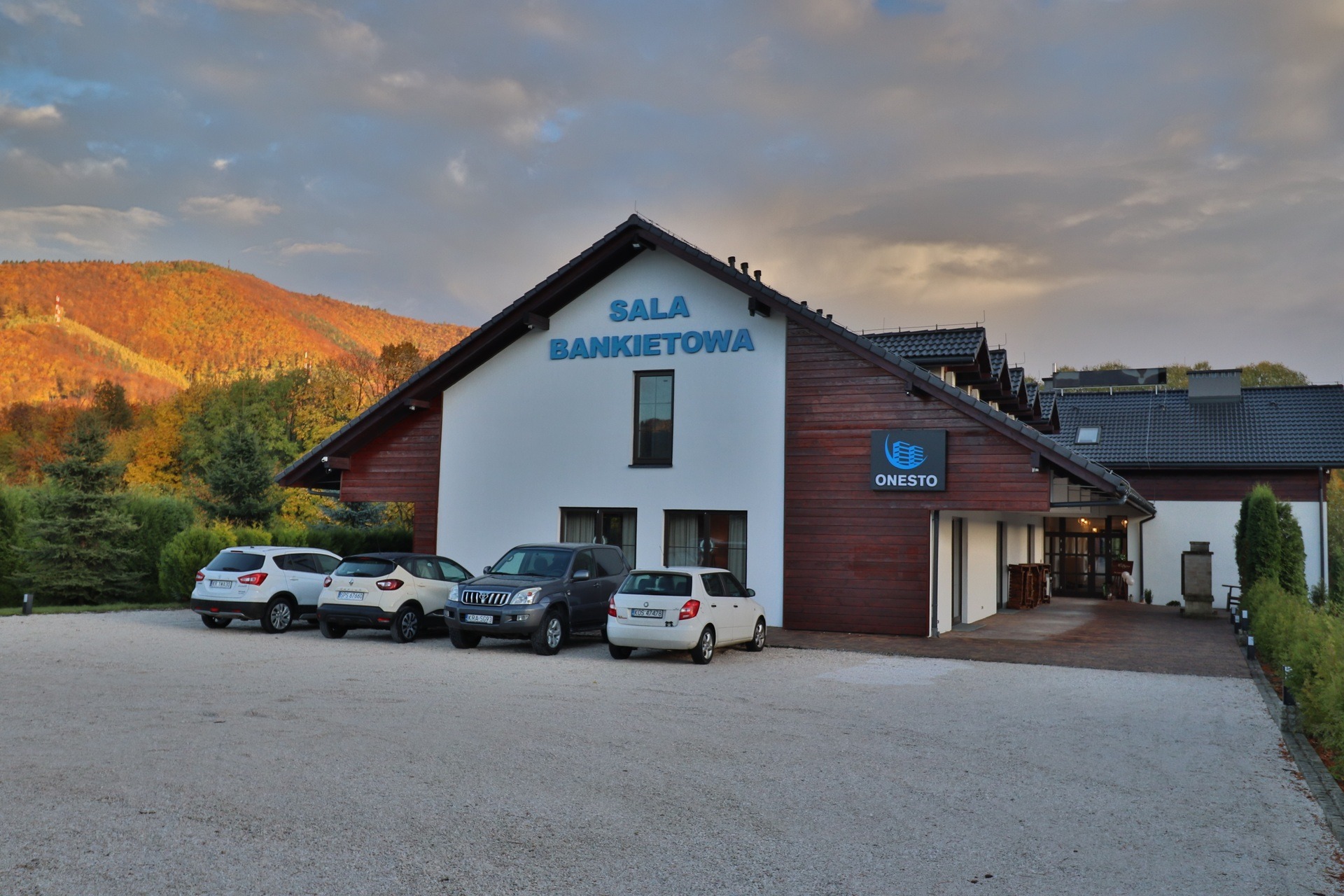 Budynek z napisem 'SALA BANKIETOWA' i logo 'ONESTO' na tle jesiennego krajobrazu górskiego oraz zaparkowane samochody.