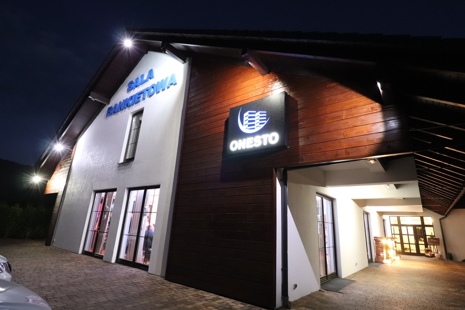 Oświetlony budynek sali bankietowej z widocznym logo firmy 'Onesto' na drewnianej elewacji, sfotografowany z perspektywy żabiej o zmierzchu.