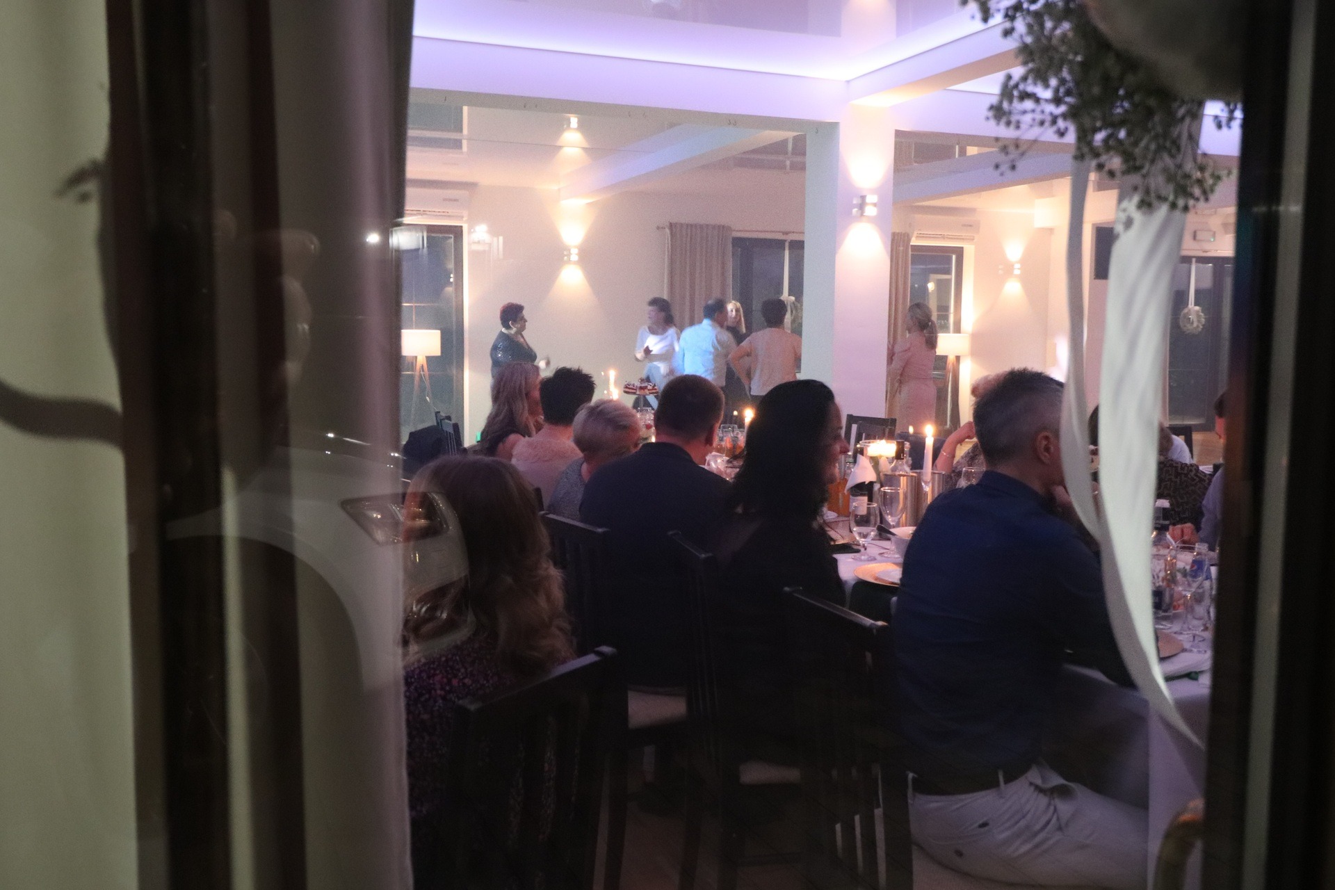 Goście siedzą przy udekorowanych stołach podczas przyjęcia w jasnym wnętrzu, widok przez okno z zasłonami.