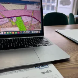 Laptop wyświetlający mapę działek na stronie swiebodzin.e-mapa.net, obok dokument z Enea Operator, na biurku w tle okno z widokiem na budynek.