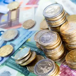 Stosy monet euro (1 i 2 euro) oraz banknoty 5, 10 i 20 euro w tle, ułożone na jasnej powierzchni.