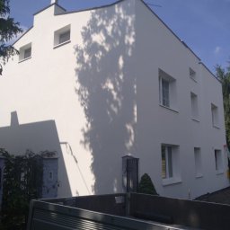 Kriss Bud Krzysztof Janiszewski - Odnowiona, biała elewacja budynku z regularnymi oknami, na której widoczne są cienie drzew. W tle błękitne niebo i fragment skrzyni ładunkowej samochodu.