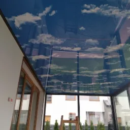 Błękitne niebo z chmurami nadrukowane na suficie napinanym w oranżerii, widoczna drabina i odbicie ogrodu w szklanych ścianach.