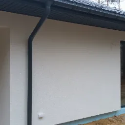 Nowo otynkowana elewacja domu jednorodzinnego z grafitowymi oknami i rynnami, widoczna warstwa izolacji fundamentów, teren wokół domu jeszcze niezagospodarowany.