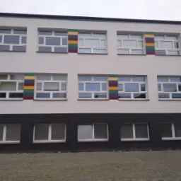 Dwukondygnacyjny budynek pomalowany na biało z czarnym pasem na dole, okna z kolorowymi paskami po bokach, widok z zewnątrz na elewację po renowacji.