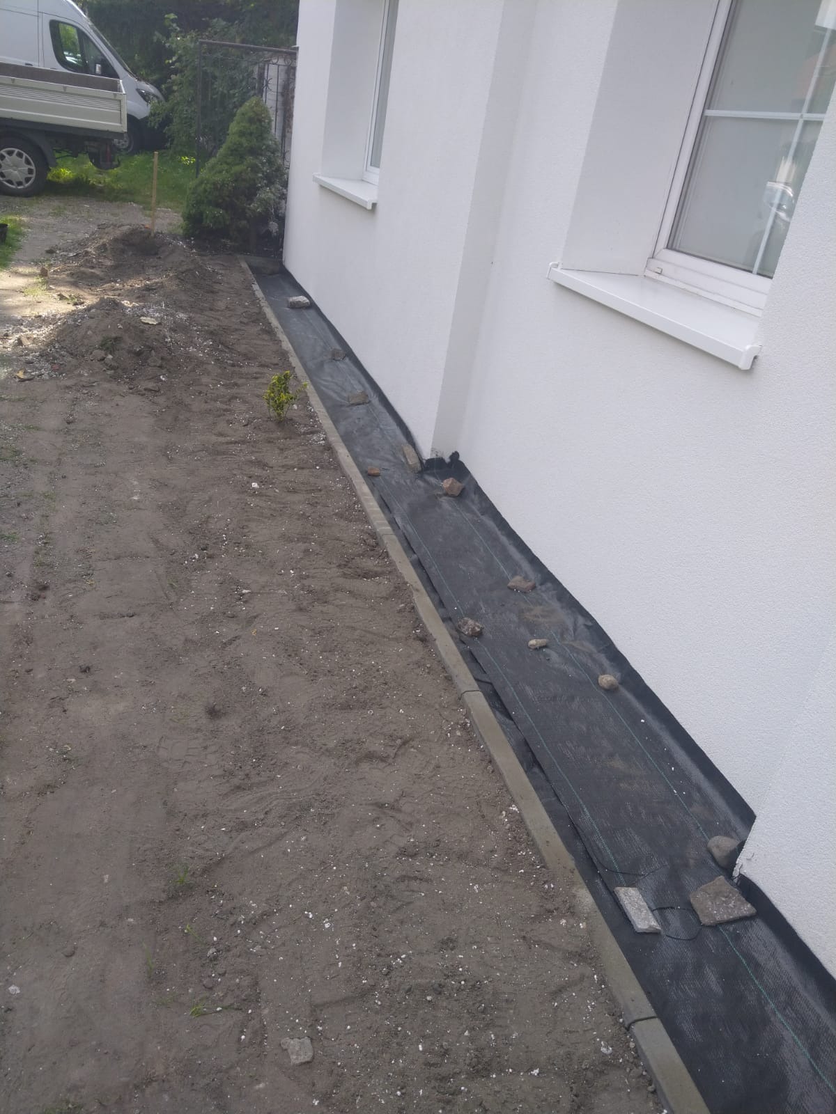Przygotowanie terenu przy ocieplonej ścianie budynku, widoczna geowłóknina zabezpieczająca grunt, obrzeża chodnikowe i świeżo otynkowana elewacja z białymi ramami okiennymi.