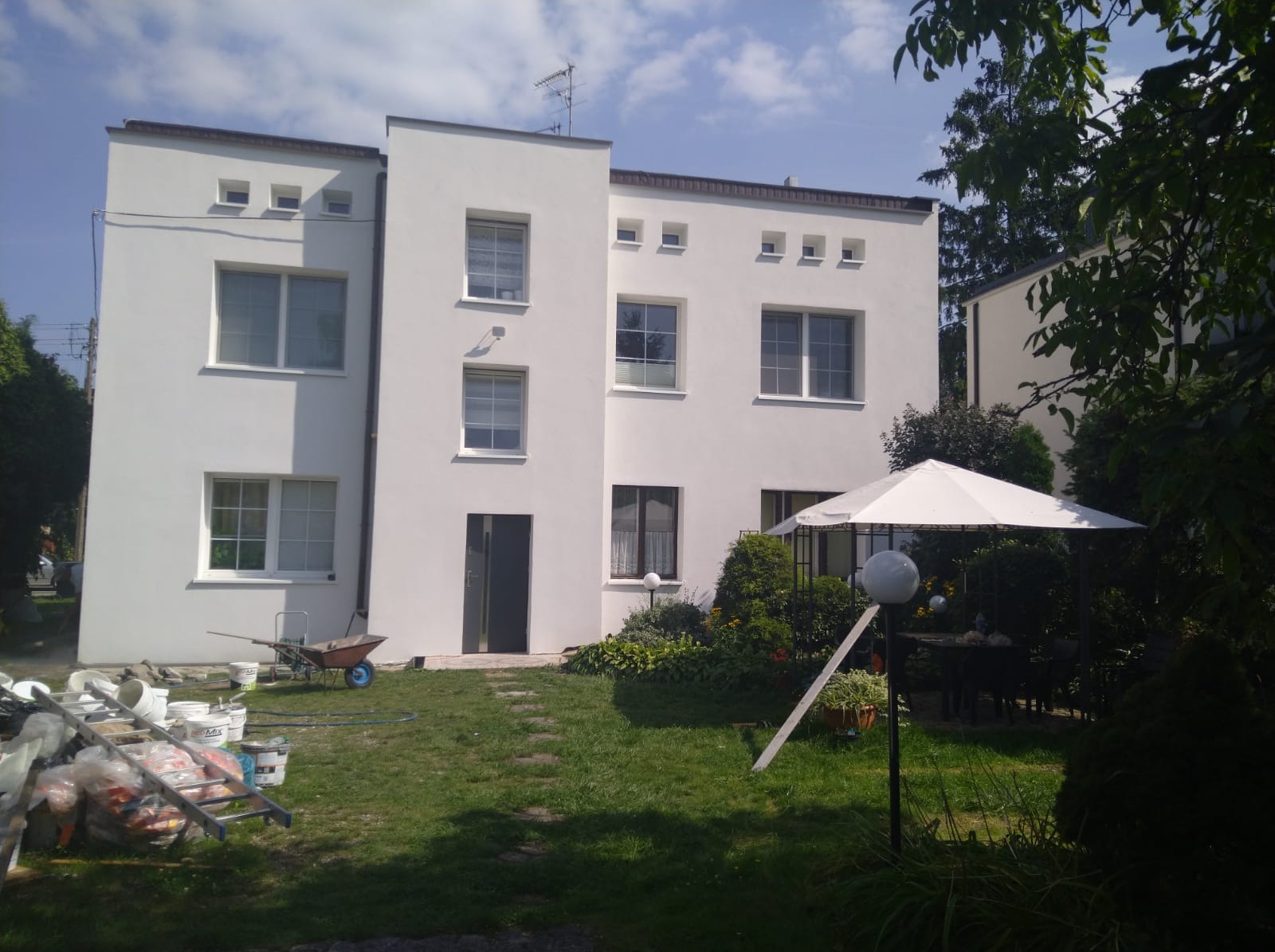 Dwupiętrowy budynek z białymi ścianami po remoncie elewacji, widoczne narzędzia i materiały budowlane na trawniku, w tle altana ogrodowa.