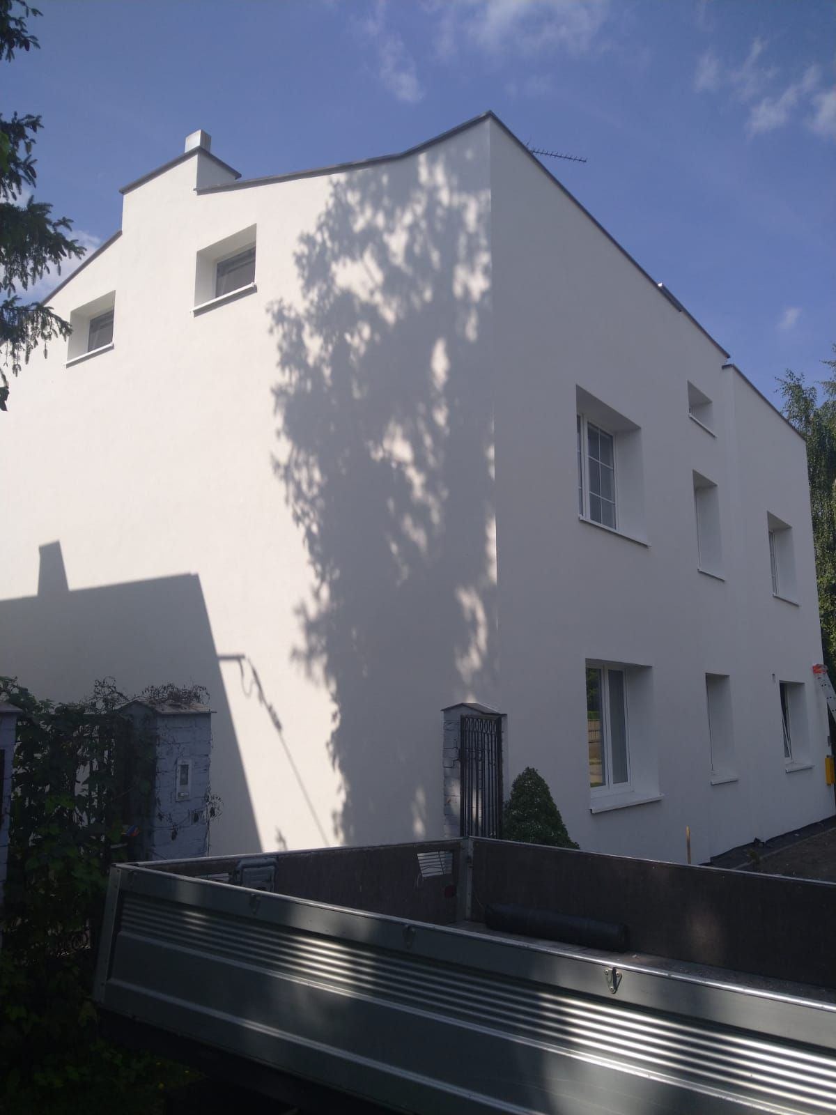 Odnowiona, biała elewacja budynku z regularnymi oknami, na której widoczne są cienie drzew. W tle błękitne niebo i fragment skrzyni ładunkowej samochodu.