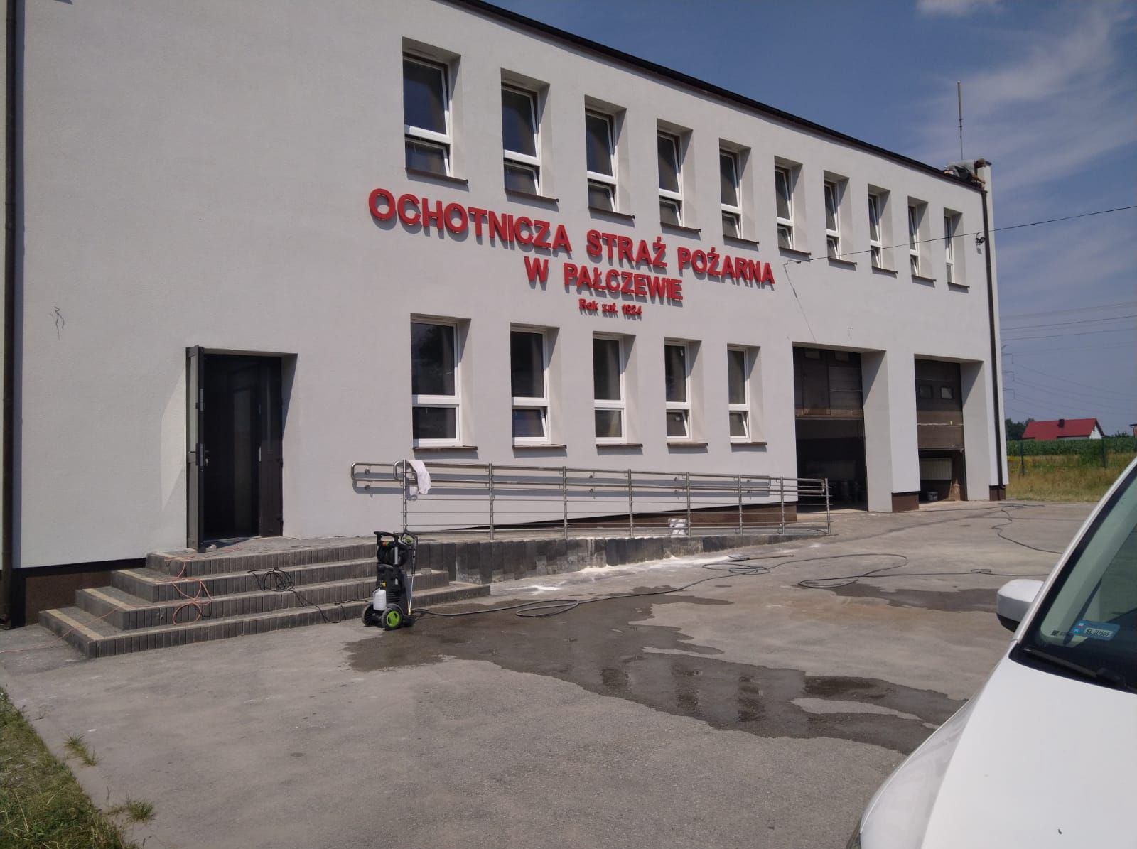 Biała elewacja budynku Ochotniczej Straży Pożarnej w trakcie czyszczenia myjką ciśnieniową, widoczne mokre plamy na chodniku, schody wejściowe i fragment samochodu.