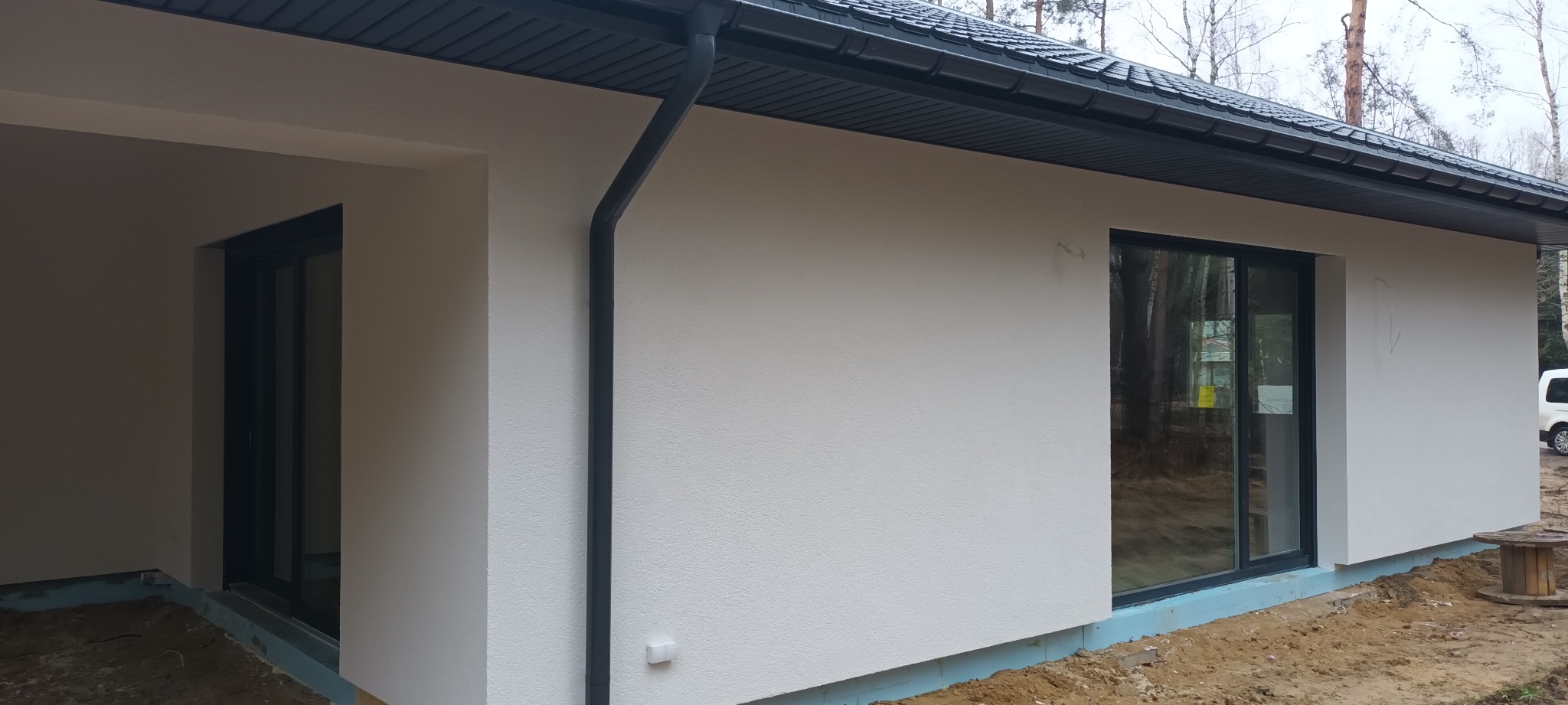 Nowo otynkowana elewacja domu jednorodzinnego z grafitowymi oknami i rynnami, widoczna warstwa izolacji fundamentów, teren wokół domu jeszcze niezagospodarowany.