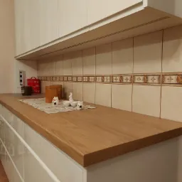 Jasny blat drewniany w kuchni z białymi, błyszczącymi frontami szafek i dekoracyjnymi płytkami na ścianie, uzupełniony ceramicznymi figurkami kotów i zajączka na haftowanej serwetce.