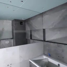 Łazienka w trakcie wykańczania: szare płytki na ścianach, częściowo zamontowane, sufit z płyt karton-gips, widoczne otwory na oświetlenie, odbicie mężczyzny robiącego zdjęcie telefonem w lustrze.