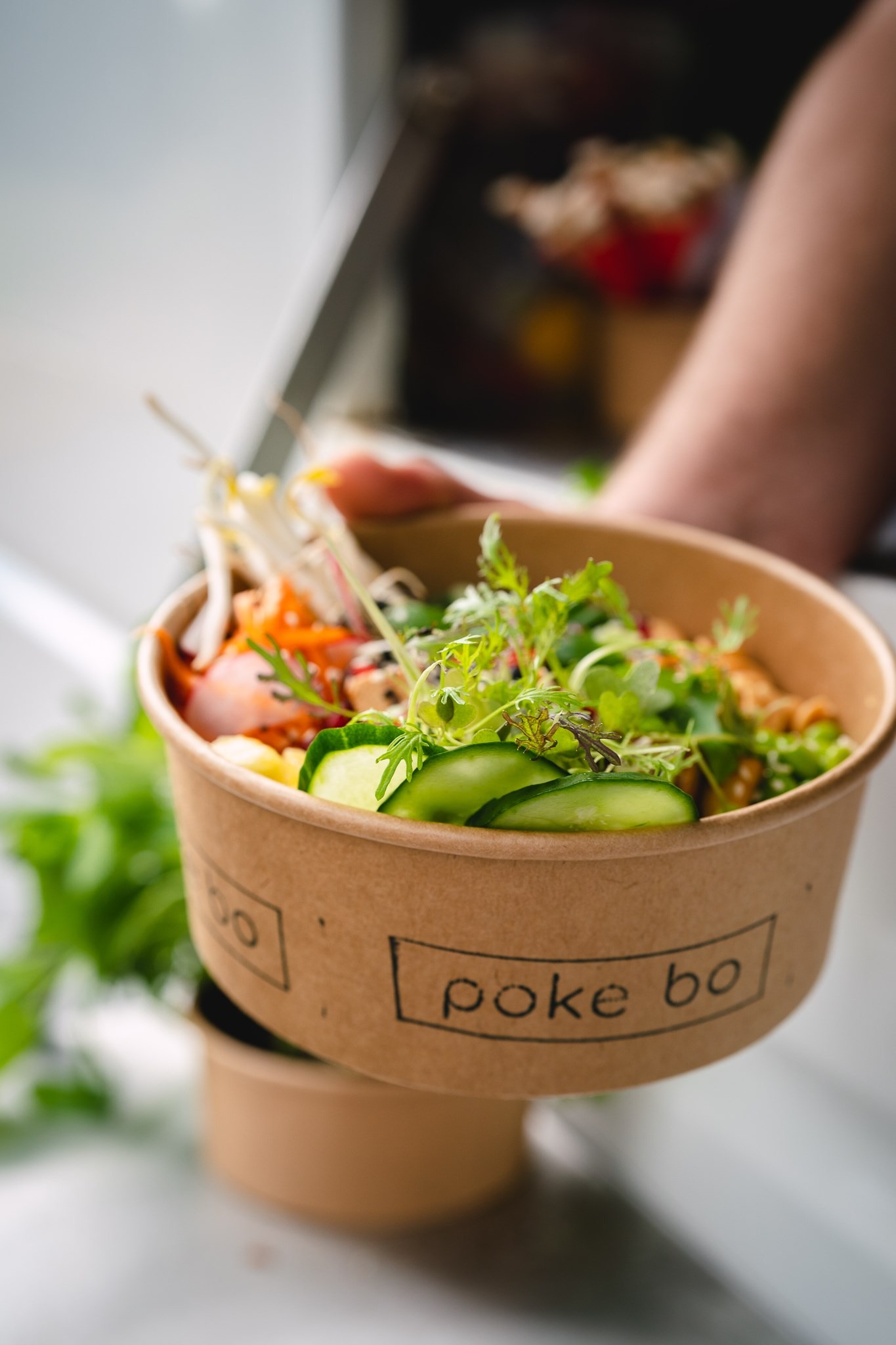 Zbliżenie na papierową miskę z poke bowl, z widocznymi świeżymi ogórkami, kiełkami fasoli mung, mikro-zielenią i innymi składnikami; w tle niewyraźna zieleń i ręka trzymająca miskę.