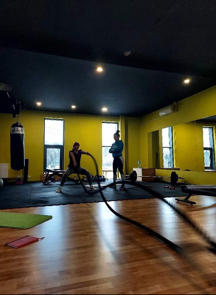 Kobieta ćwiczy z linami w siłowni, druga osoba obserwuje, w tle worki bokserskie i sprzęt do ćwiczeń.